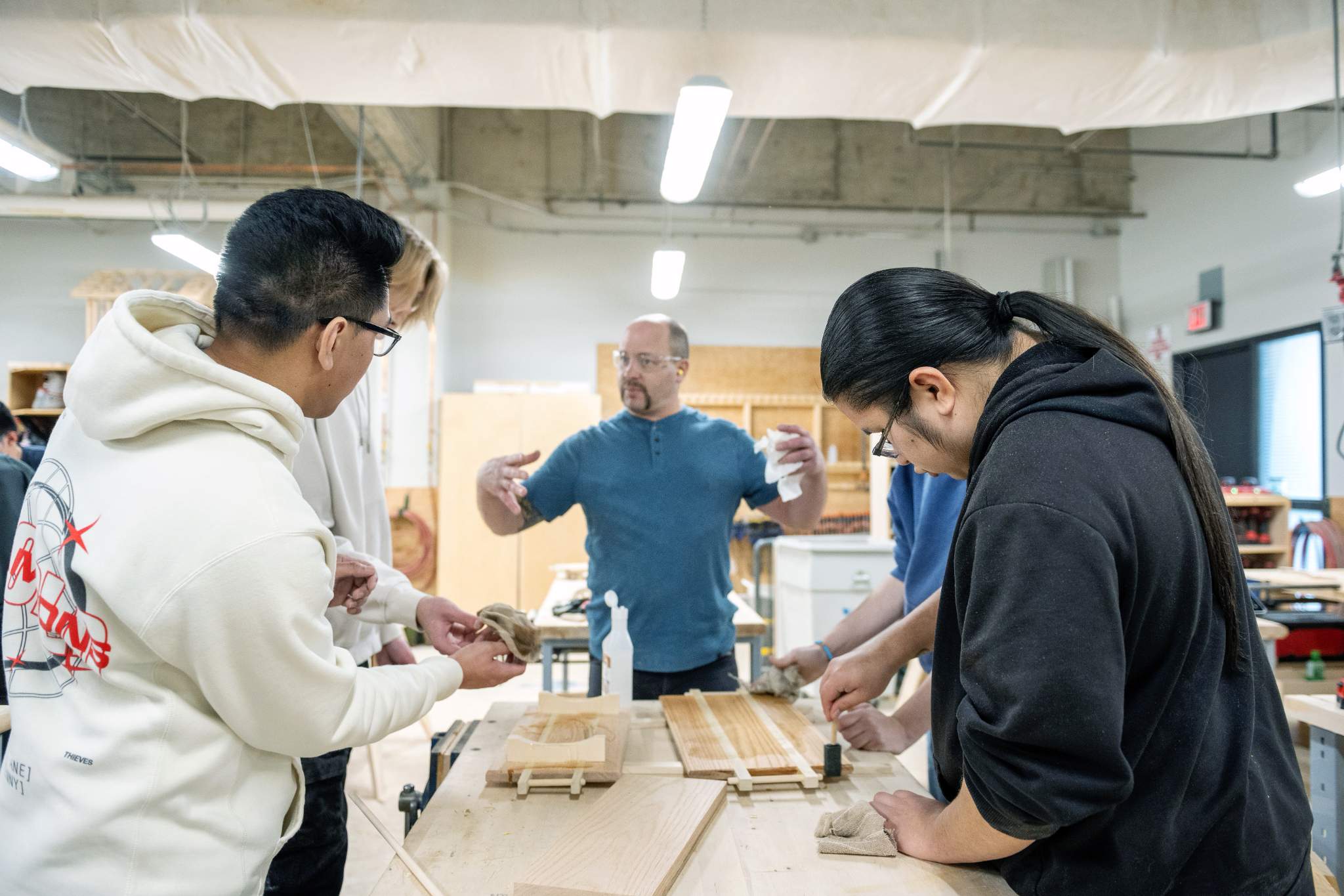 A carpentry instructor demonstrates a woodworking technique to two youths wearing safety glasses in a workshop, with tools and materials visible on a workbench.