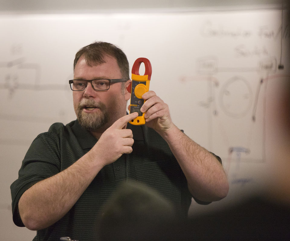 An instructor demonstrating the use of a clamp meter while explaining electrical circuit concepts.
