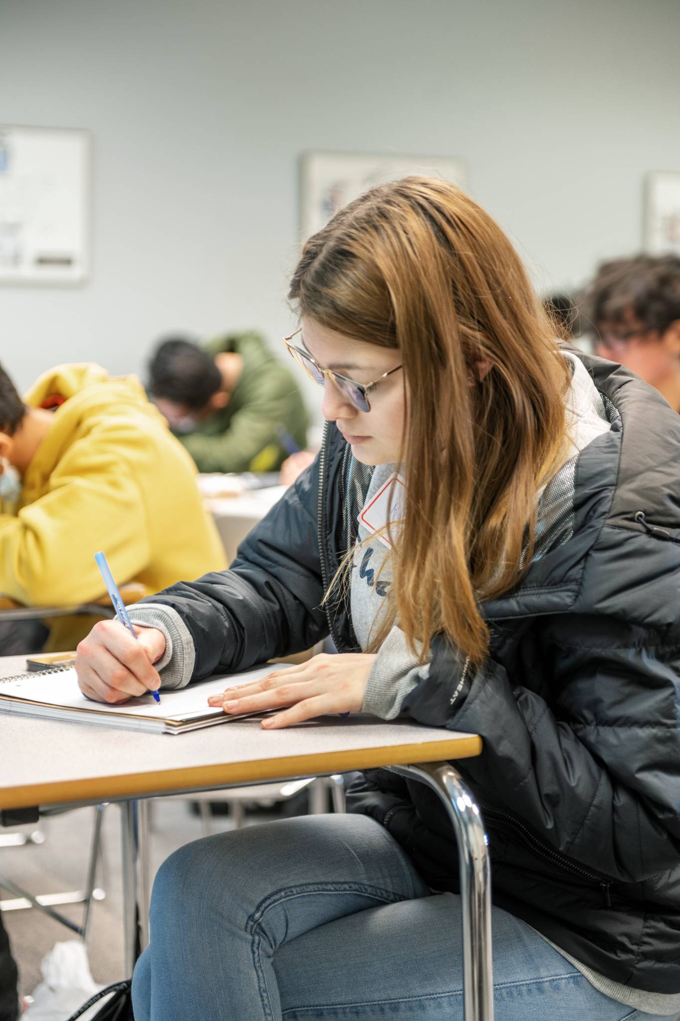 Students wearing safety glasses work on hands-on projects at a workshop table, using tools and measuring materials while others work in the background.
