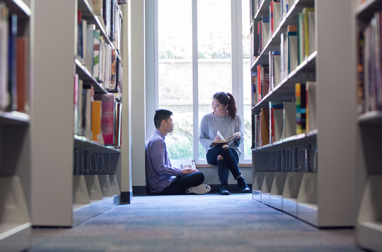 Two students sit on the floor between library bookshelves, talking and studying near a large window.