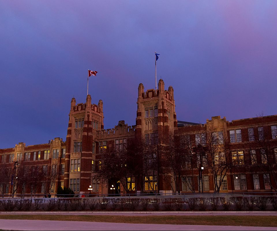A wide view of a historic brick academic building with twin towers and flags on top.