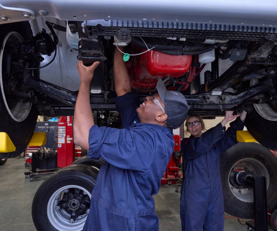 Two students working beneath an elevated vehicle in an automotive shop.