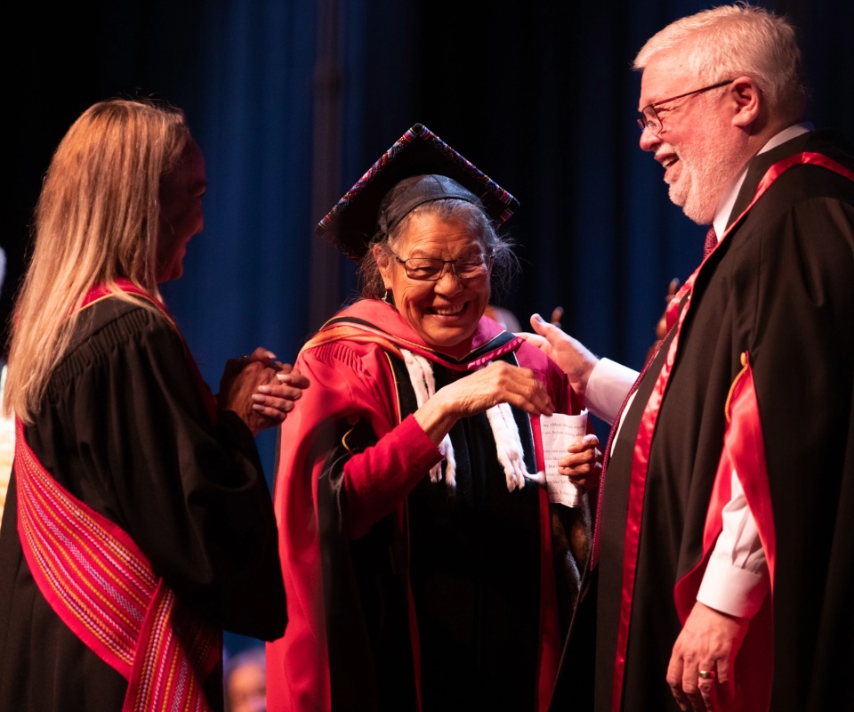 A joyful honourary degree recipient in academic regalia smiles and shakes hands with an official while another attendee claps in celebration.