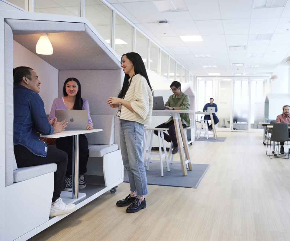 An interior office scene featuring a collaborative workspace with individuals using laptops in a cozy booth and standing desks, promoting teamwork and modern design.