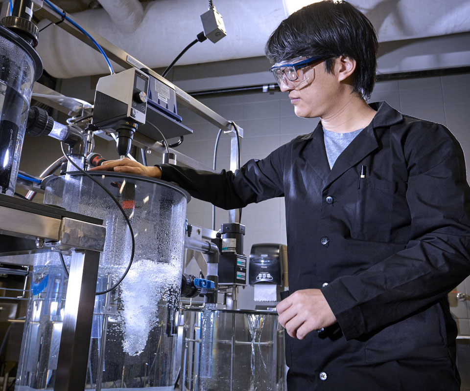A student researcher in a lab coat conducts an experiment with a large glass container filled with liquid, using scientific equipment.