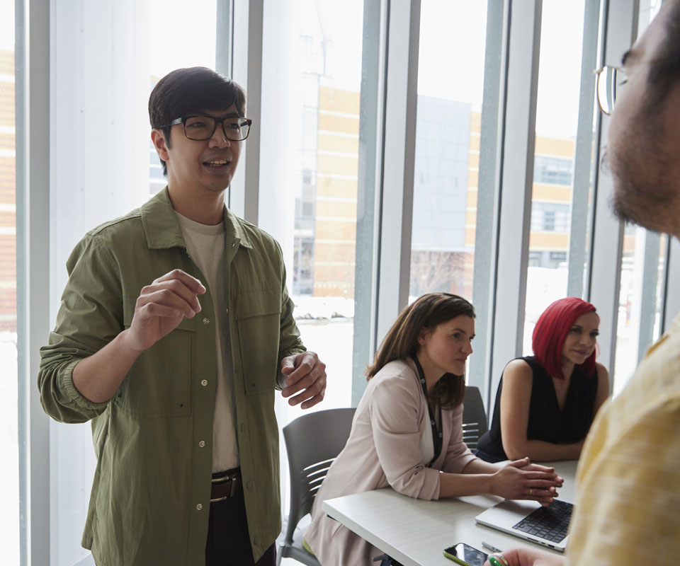 A young man in a green jacket engages in a discussion with colleagues at a modern office setting. Two women seated at the table appear to be listening attentively.