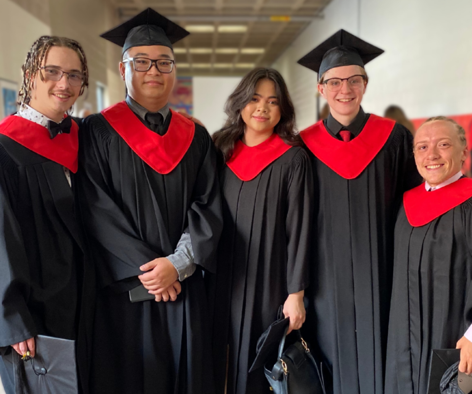 Group of five graduates in black gowns and red collars, smiling together in a school hallway.