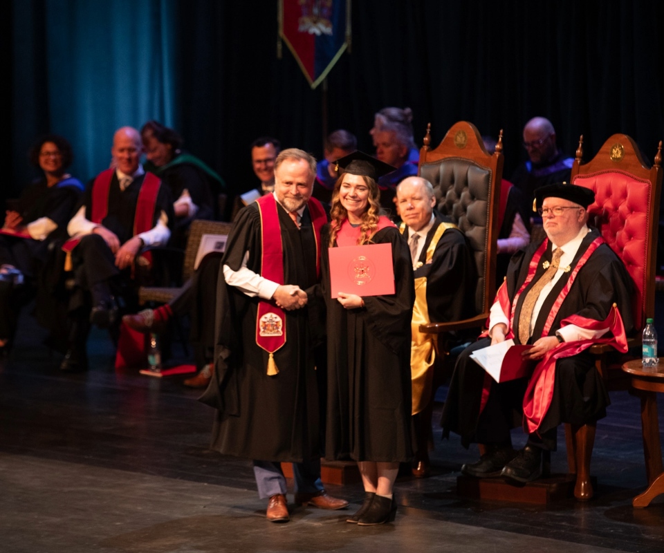 A graduate receiving their diploma on stage during a graduation ceremony, surrounded by faculty members in traditional academic regalia.