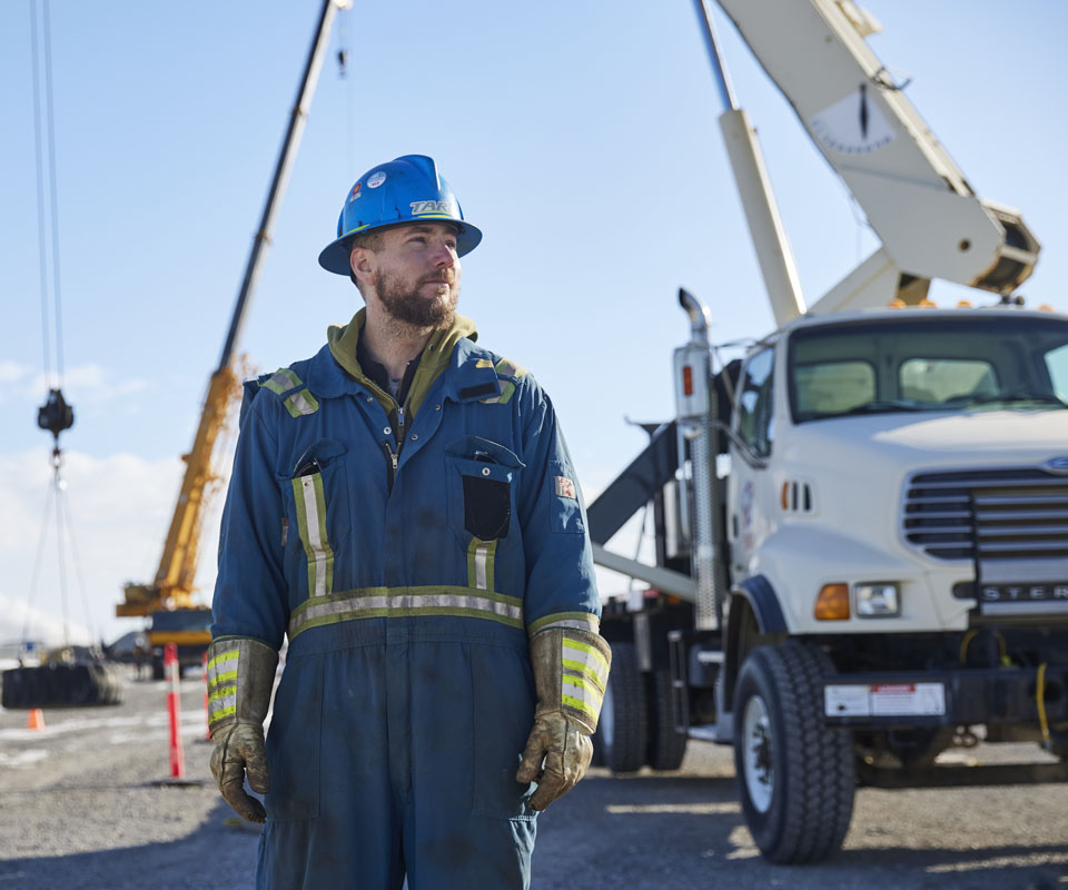 A crane operator apprentice in a hard hat stands confidently near a crane and a truck on a worksite.
