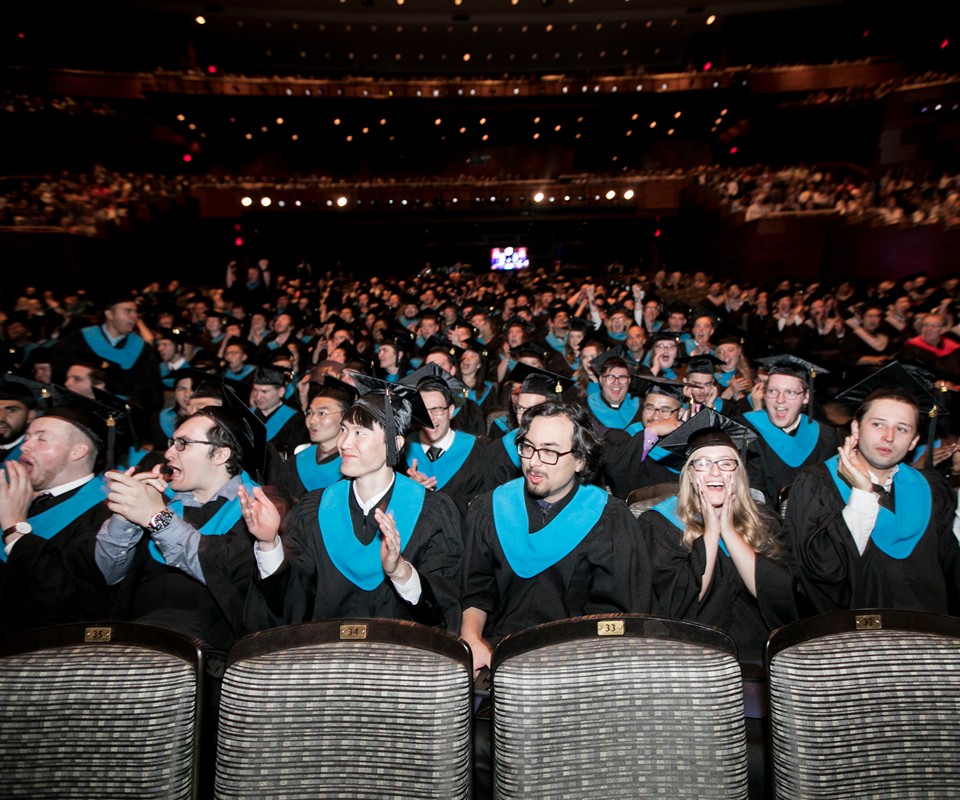 A large audience of graduates in black caps and gowns, wearing turquoise stoles, clapping and celebrating at a commencement ceremony.