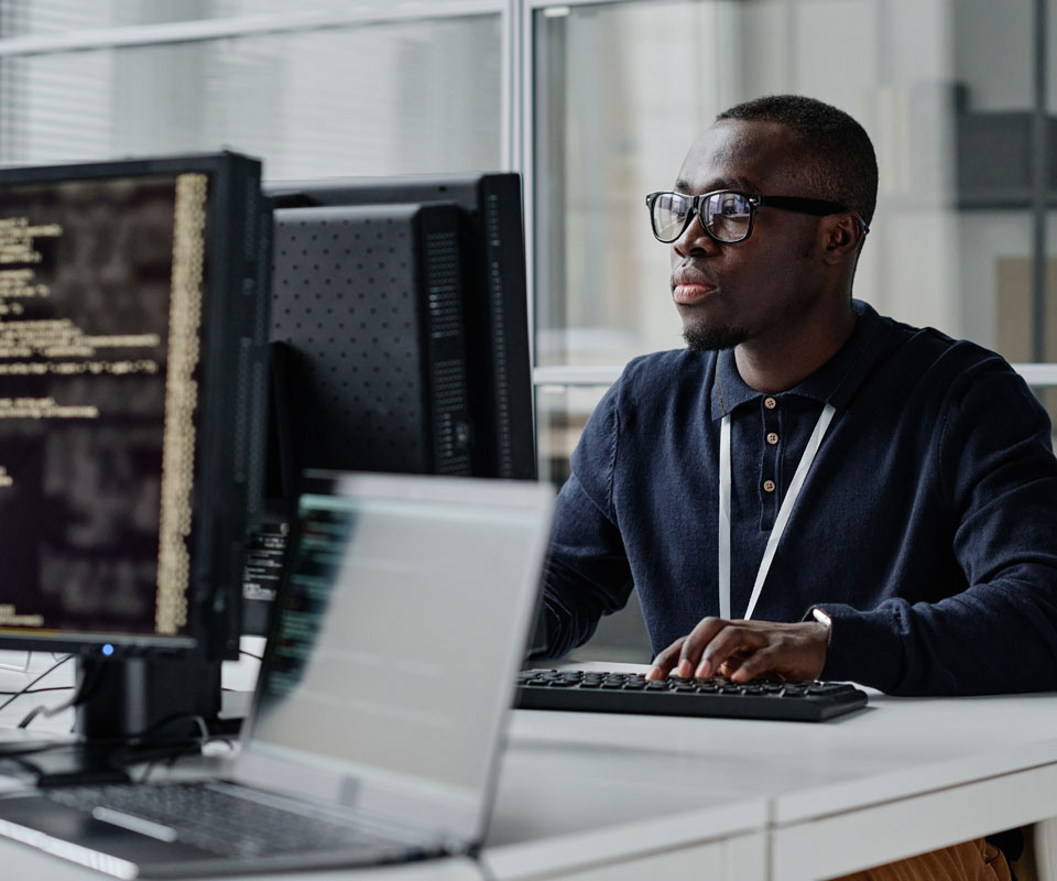 A focused young man in glasses working at a computer with code displayed on the screen.