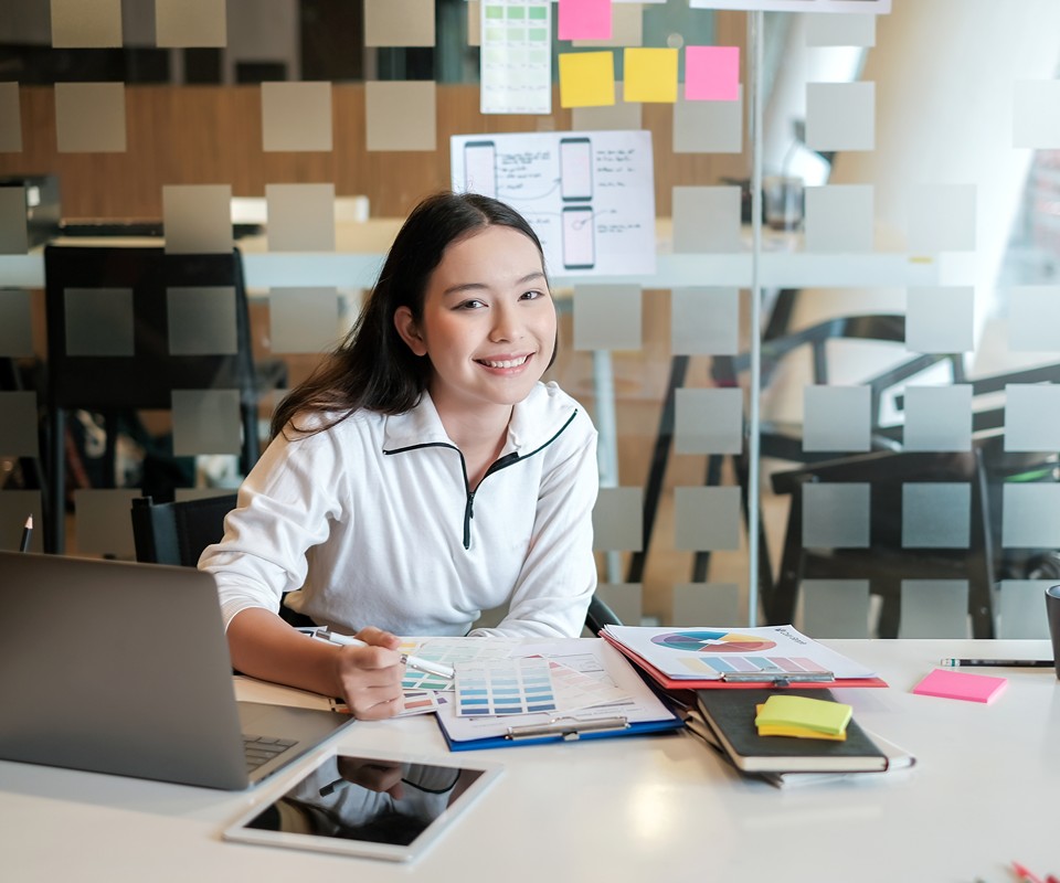 A young woman is smiling while sitting at a desk, surrounded by design materials and technology in a modern workspace.