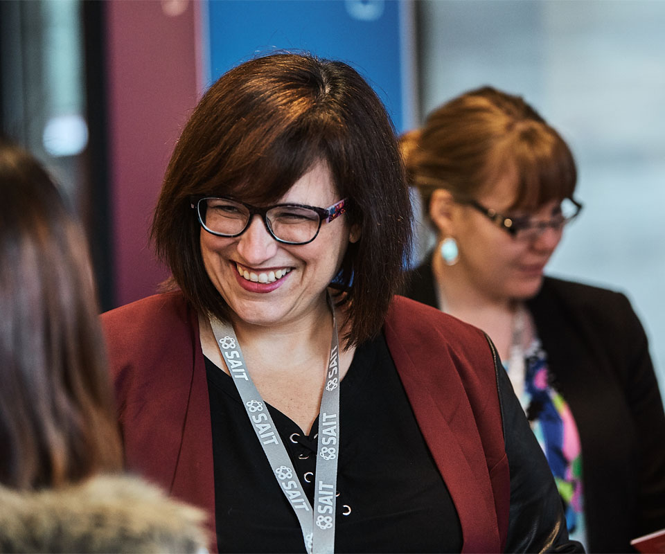 A SAIT employee with glasses and a red blazer smiles and engages in conversation at an event, with another woman in the background.