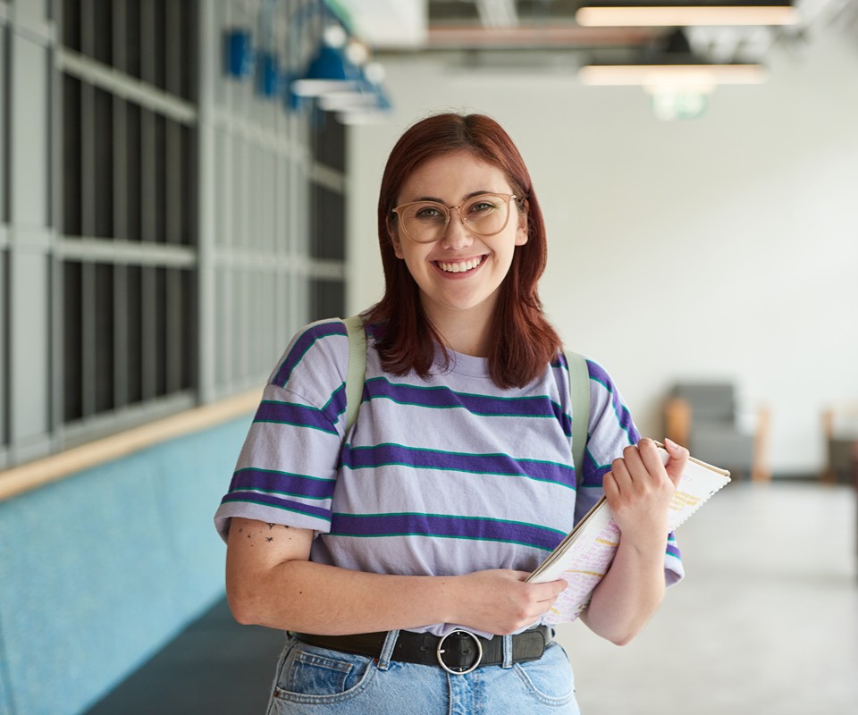 A smiling young woman in a striped shirt holds a notebook while standing in a modern interior space.