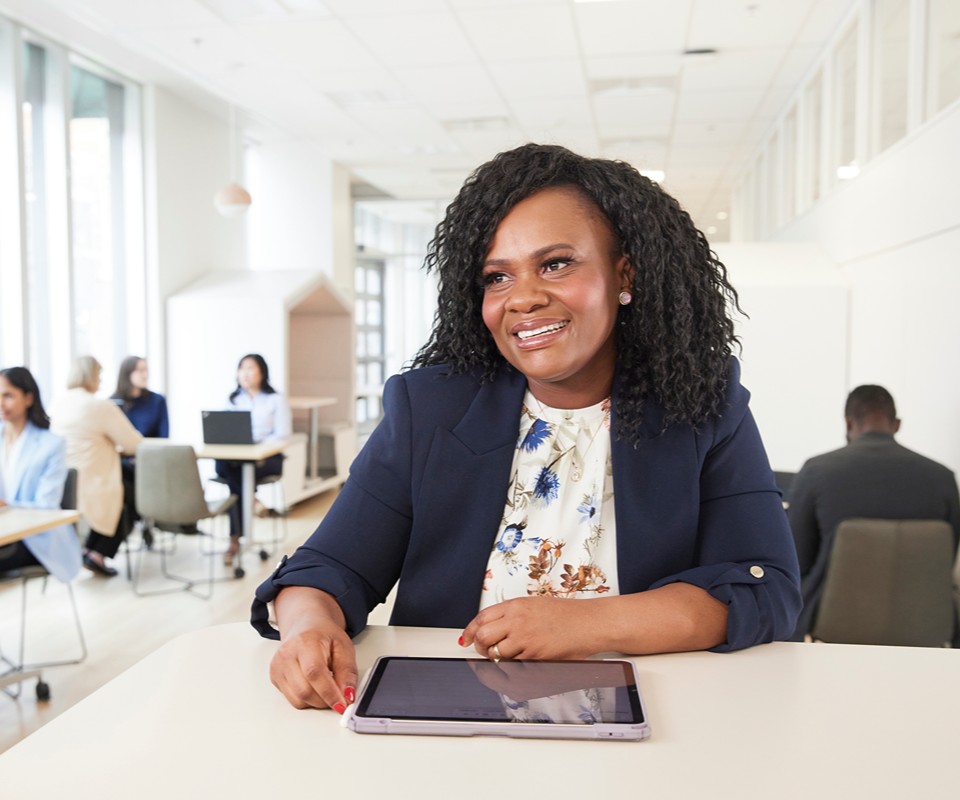A professional woman smiles while sitting at a table with a tablet in a modern office setting. The background features colleagues working collaboratively.