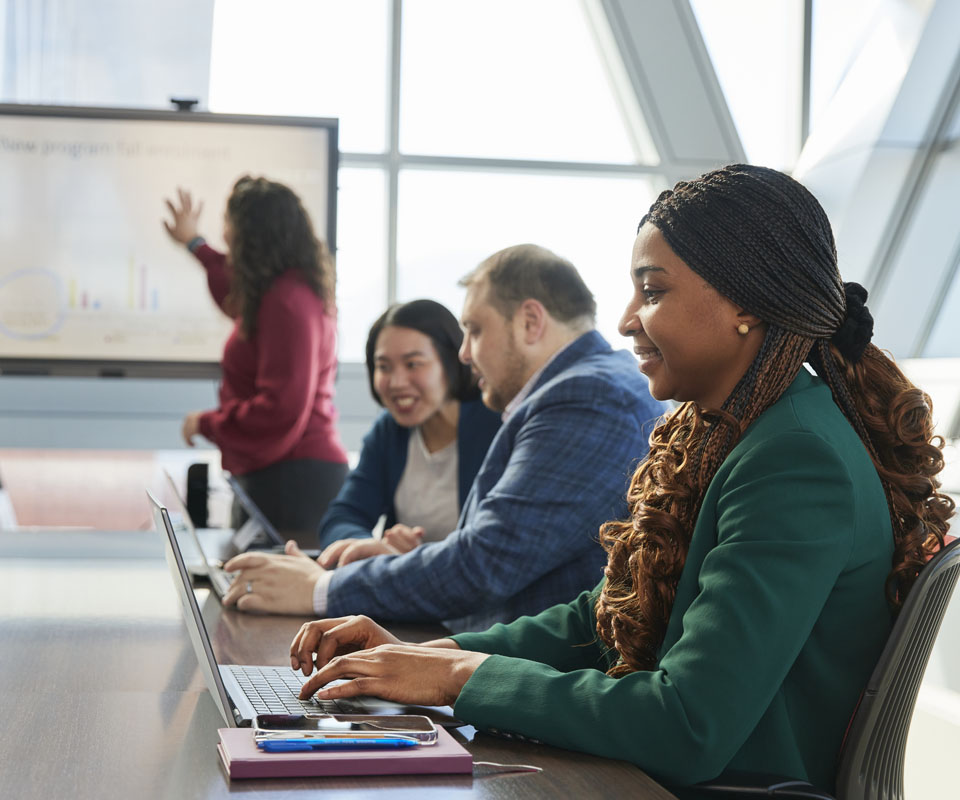 A group of professionals engaged in a meeting with a woman typing on a laptop in the foreground, while others collaborate and present ideas in the background.