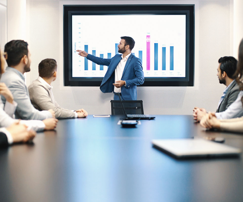 A professional presenting sales data during a business meeting, with a large screen displaying colourful graphs and charts.
