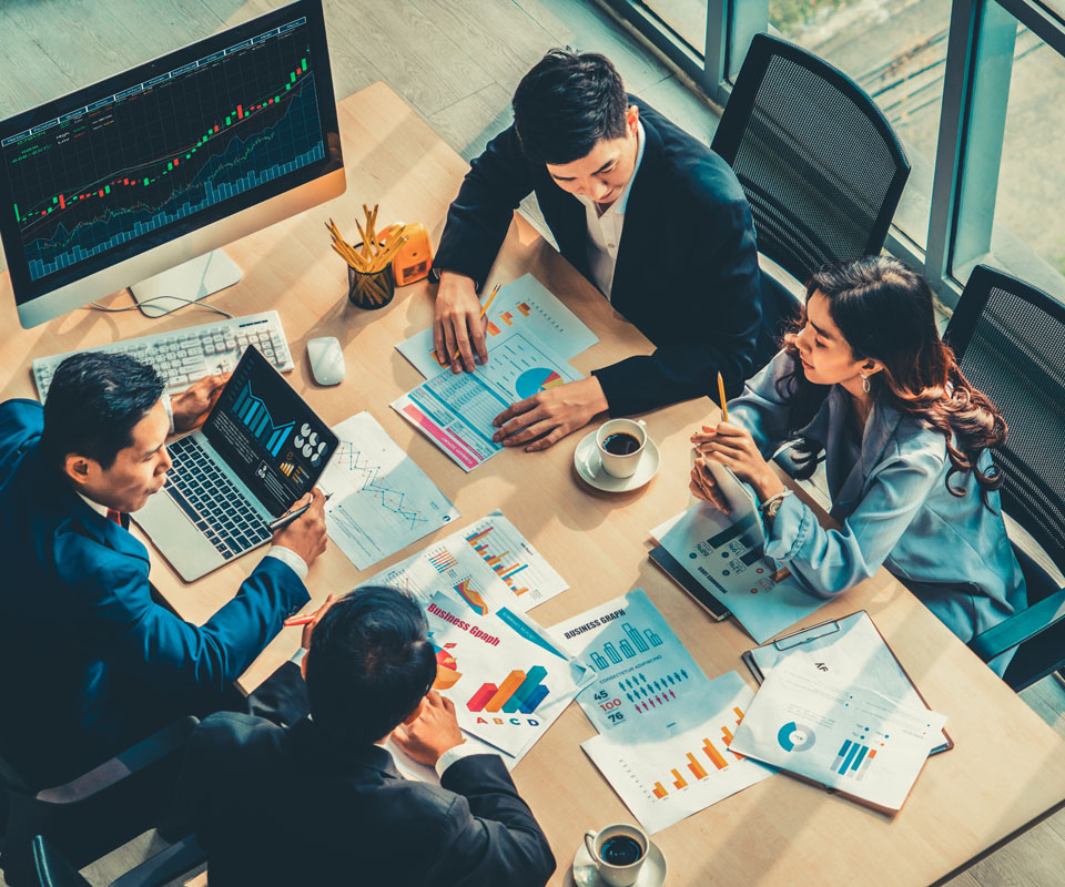 A group of four business professionals engaged in a discussion around a table filled with charts, graphs, and a laptop displaying data analytics.