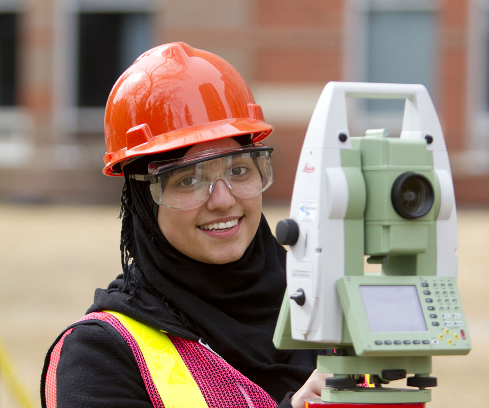 A female surveyor wearing an orange hard hat and safety vest smiles while operating a surveying instrument.