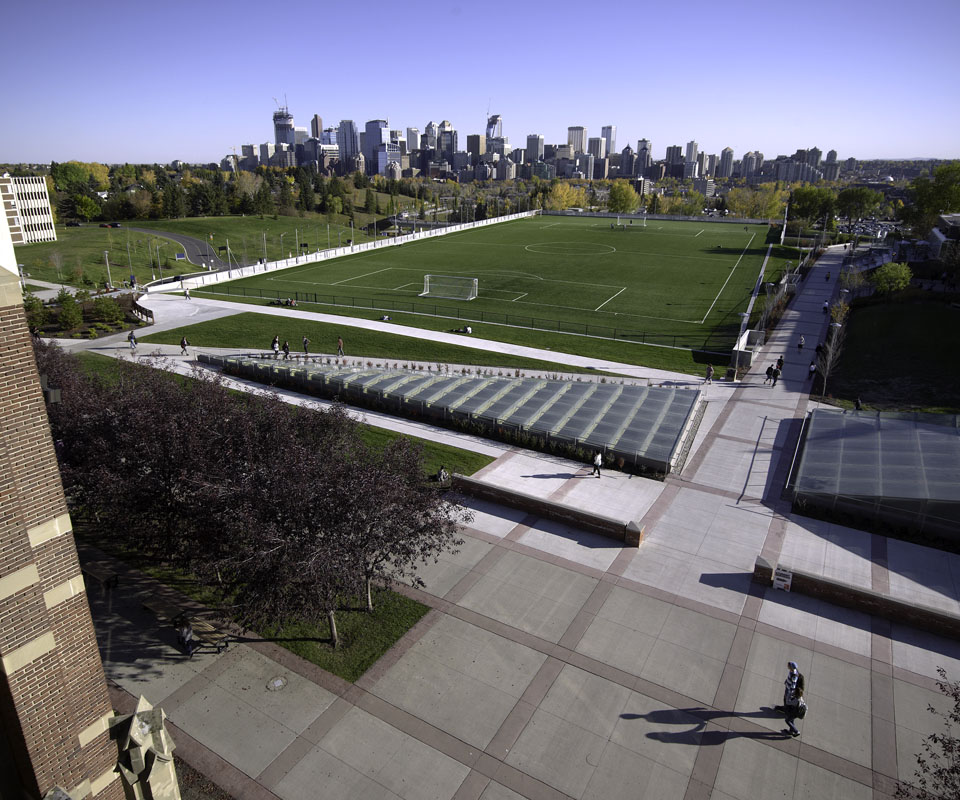A panoramic view of the southwest side of SAIT campus, featuring the Cohos Commons field and the Calgary city skyline in the background.