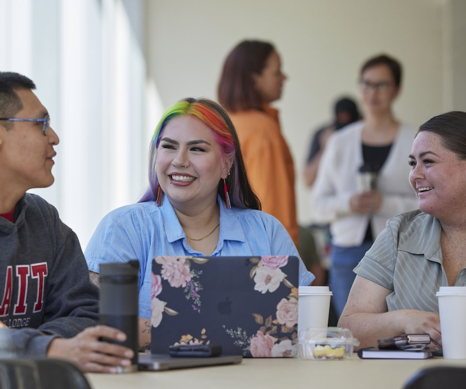 Group of diverse individuals engaged in a friendly conversation at a table with laptops and coffee cups.
