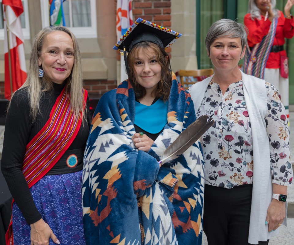 An Indigenous graduate stands between two people, wearing regalia and smiling.