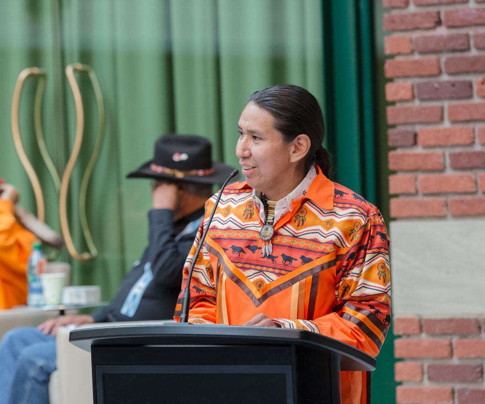 A man in a colorful traditional shirt speaks at a podium, while another person in a black hat sits in the background.