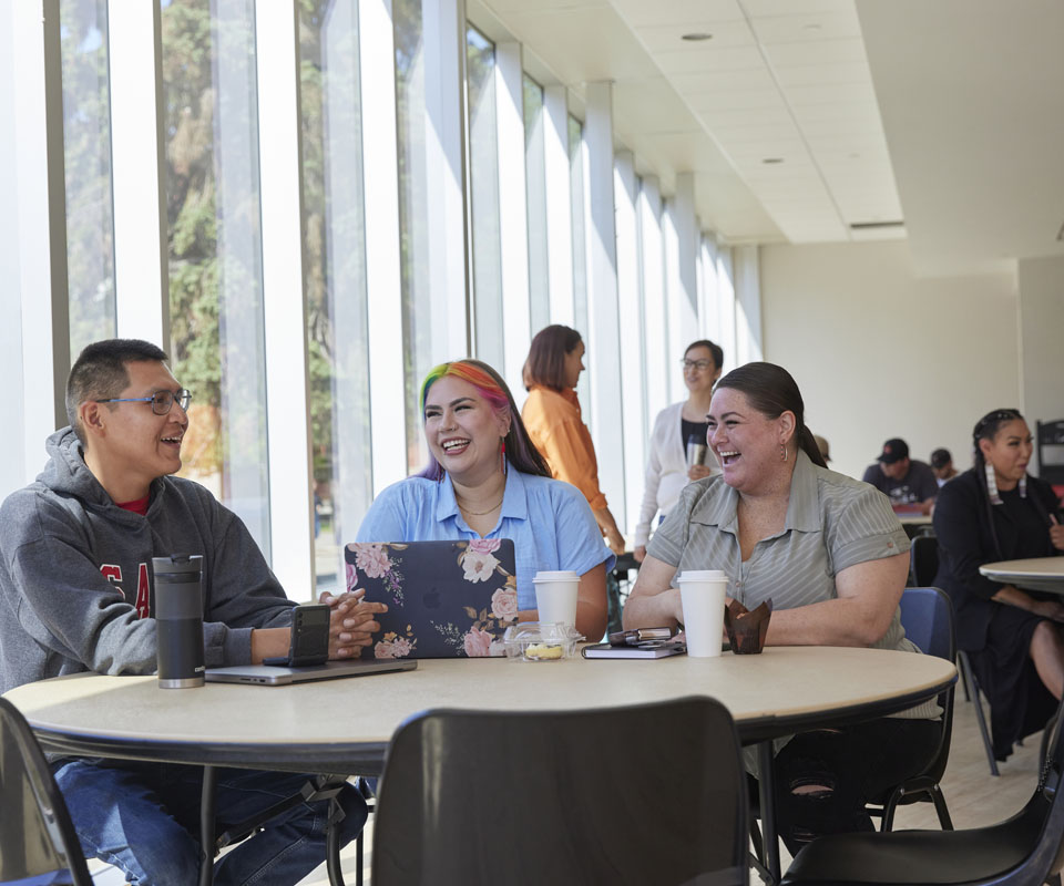 A group of three Indigenous students smiling and engaging in conversation at a table in a bright, modern space.