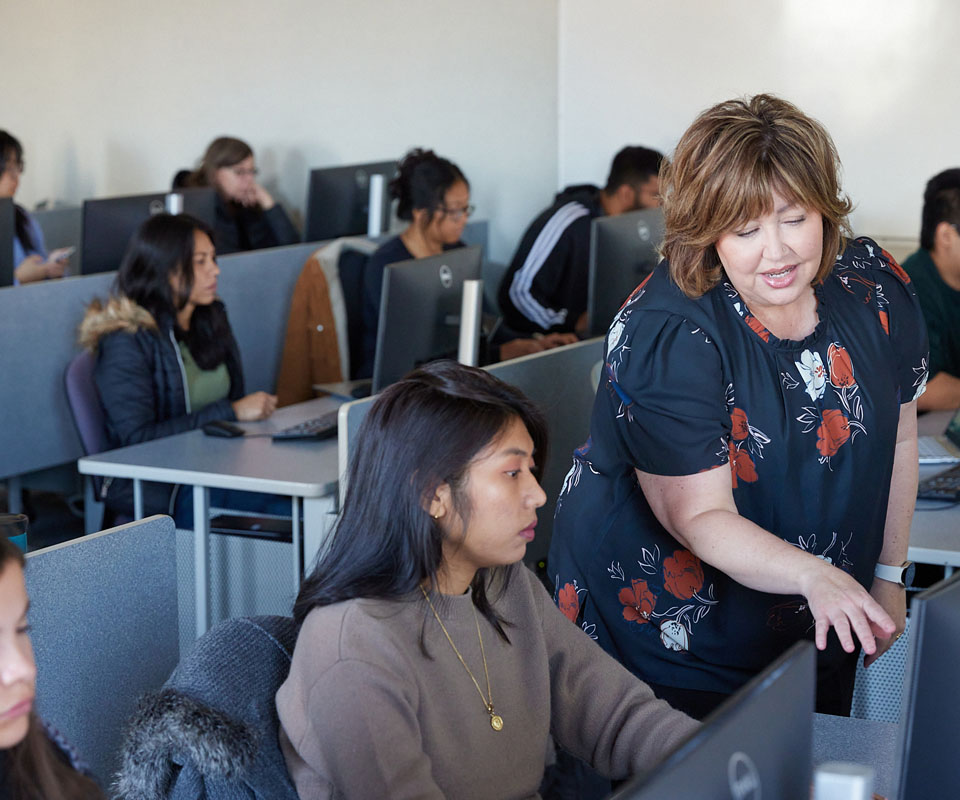 An instructor assists a student in a computer lab filled with attentive learners at their desks.