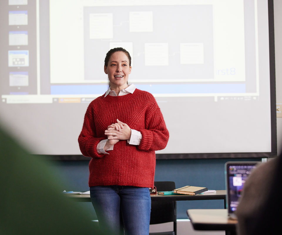 An instructor in a red sweater stands in front of a projector screen, engaging with her students during a presentation.