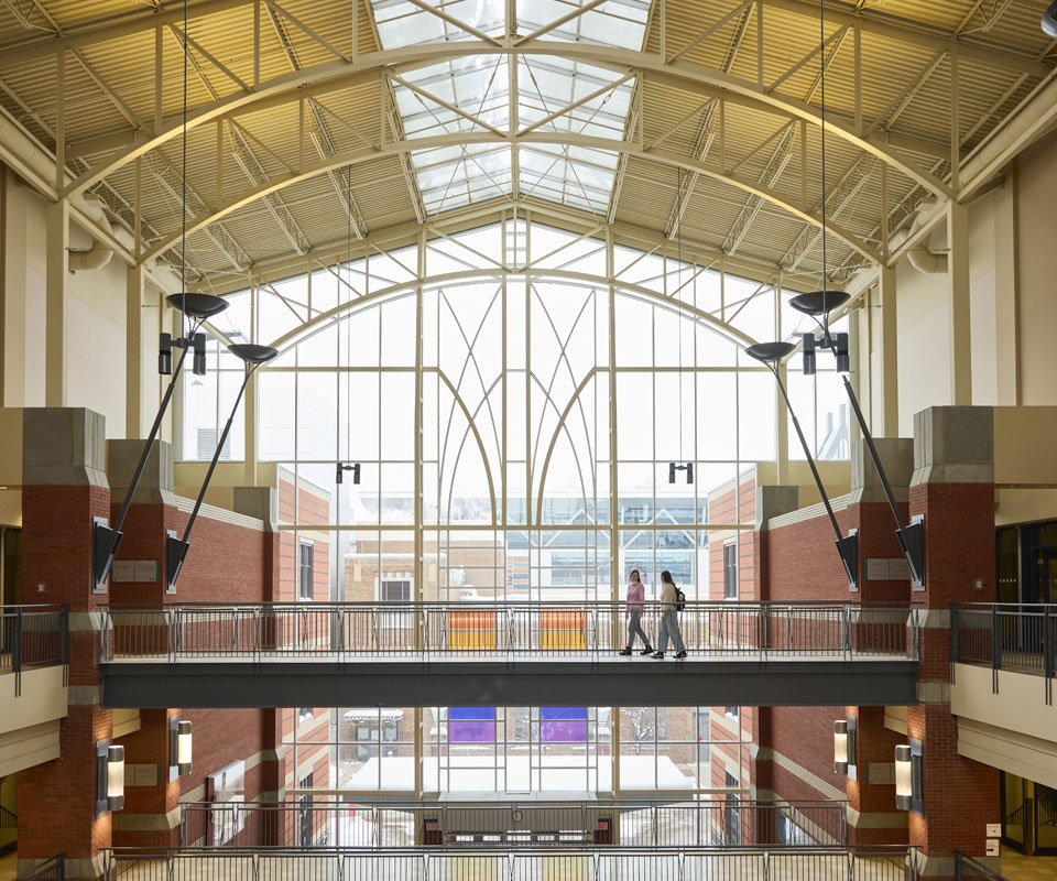 A spacious interior view of the Irene Lewis Atrium featuring a large glass roof and a bridge walkway with two people walking across it.