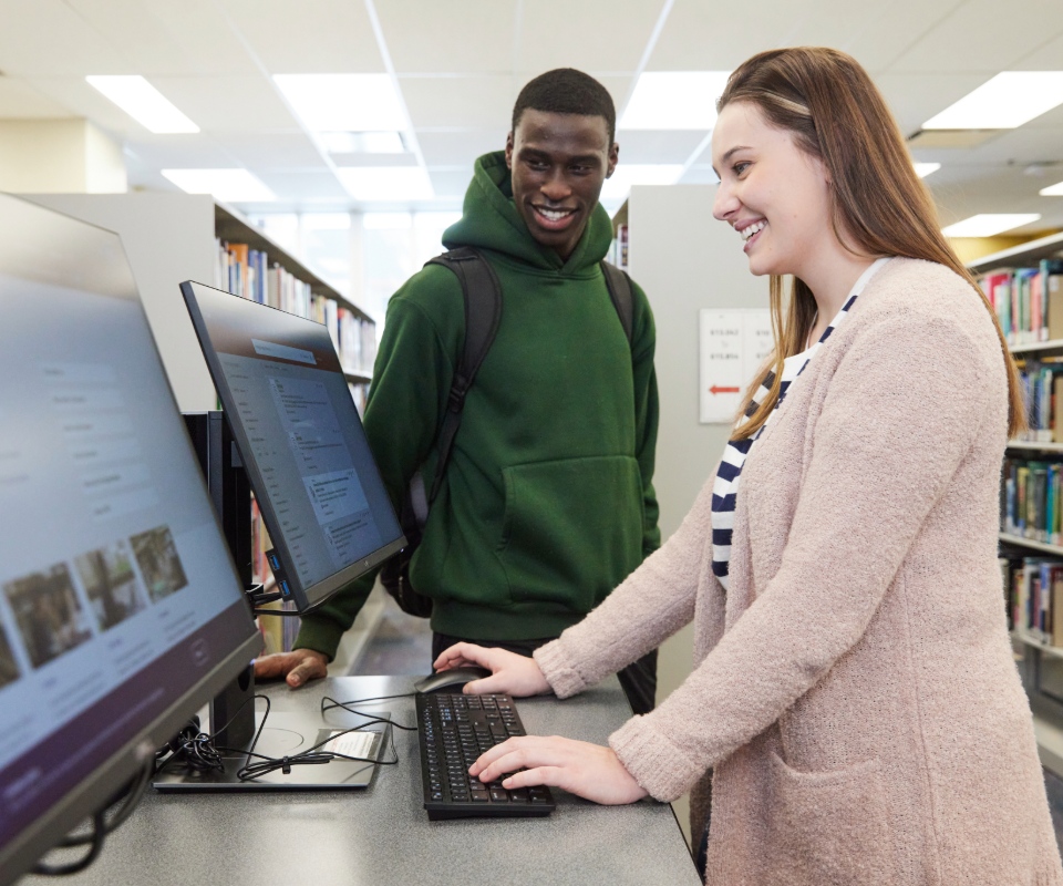 A young woman using a computer in a library while a smiling young man watches her.