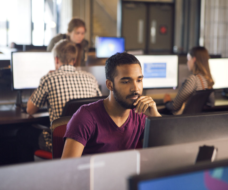 A young man focused on working at a computer in a modern computer lab, surrounded by other students engaged in similar tasks.