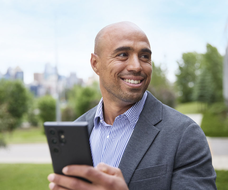 A smiling man in a blazer holds a smartphone outdoors with a city skyline in the background.