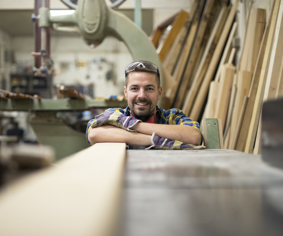 A cheerful carpentry student leaning on a workbench in a woodworking shop, surrounded by tools and raw materials.