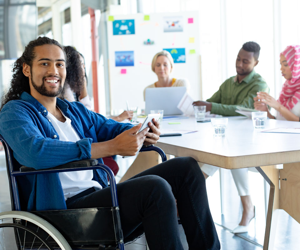 A smiling man in a wheelchair holds a phone while participating in a meeting with a diverse group of people.