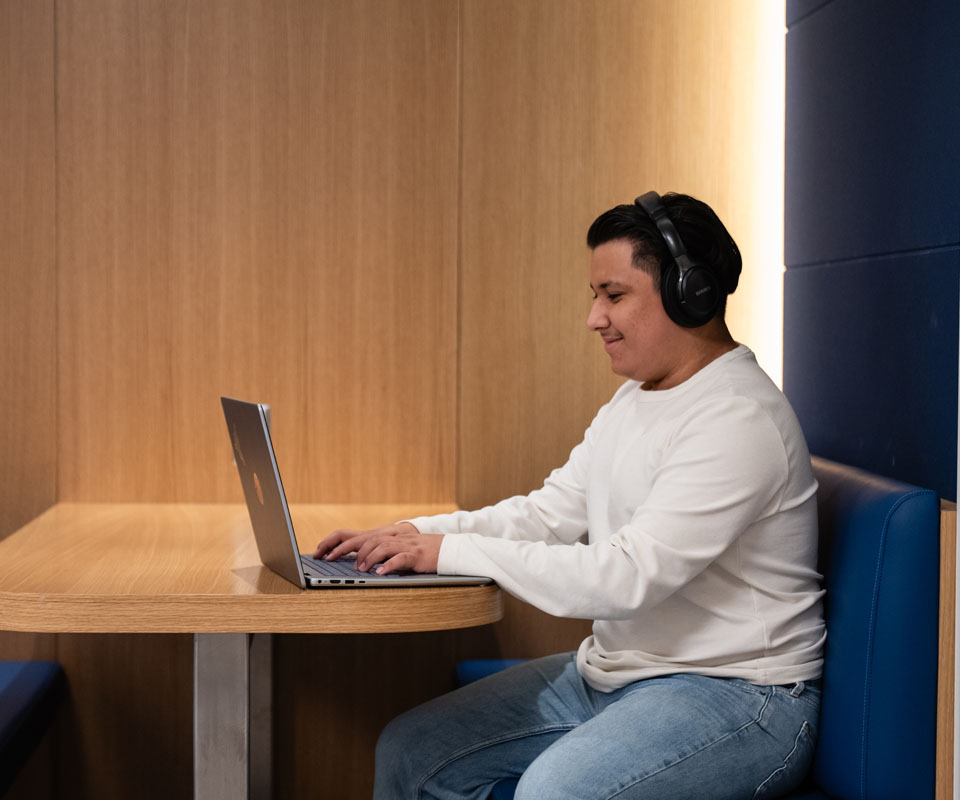 A student wearing headphones sits at a table, focused on a laptop.