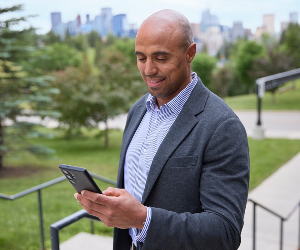 A man in a blazer smiles while using a smartphone outdoors, with a city skyline in the background.