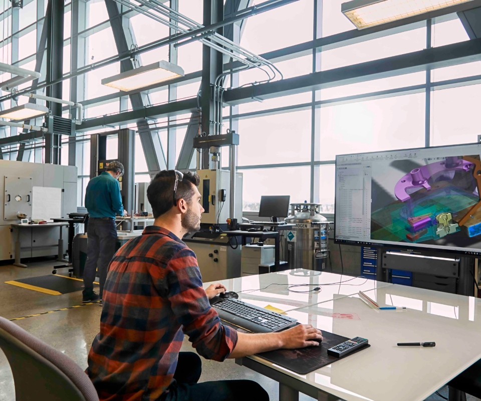 A technician working on a computer in a modern workshop with large windows, while another person operates equipment in the background.