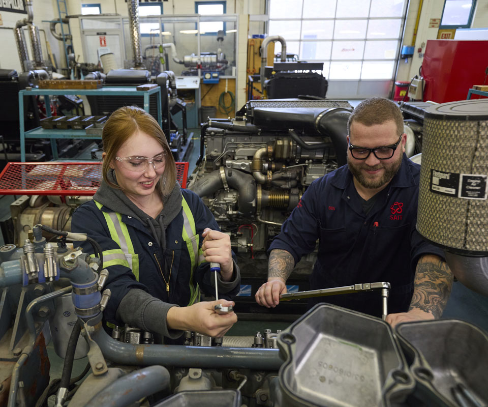 Two students working on an engine in a workshop, one is a woman in safety glasses and a work jacket using a tool, while the other, a man in a dark shirt, assists her.