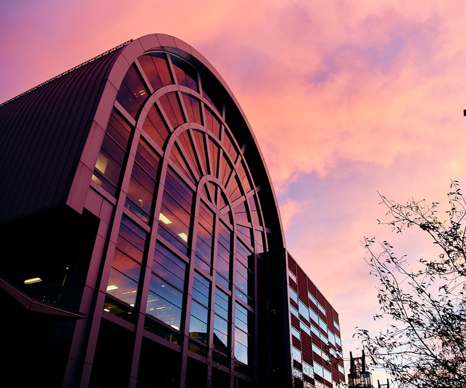 A modern building with a tall, arched glass and curved roofline, photographed from below at sunset.