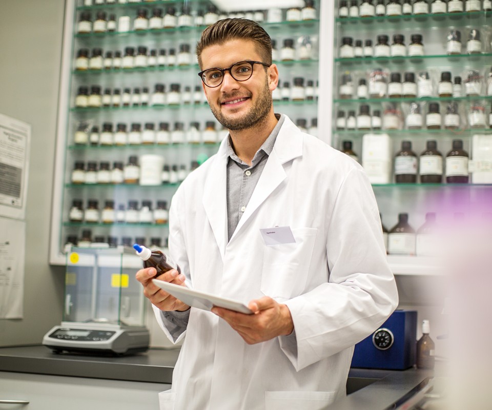 A laboratory technician in a white coat holding a clipboard and dropper, smiling in a well-stocked lab.