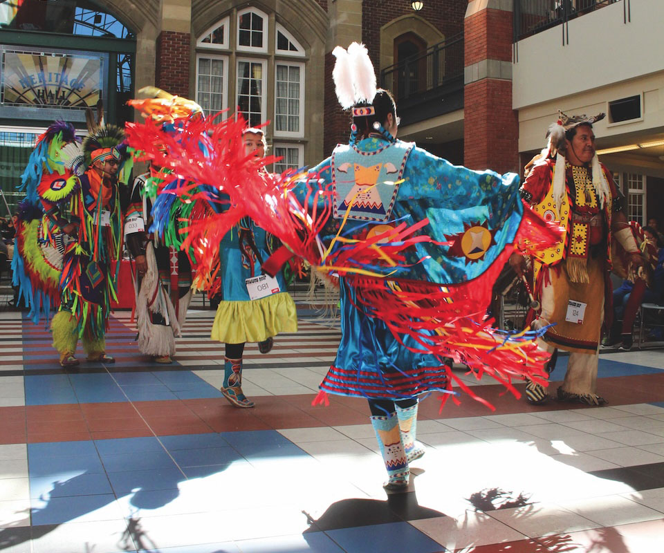 A group of dancers in colourful traditional Indigenous attire performing at a cultural event. Their vibrant costumes feature intricate patterns and feathers, showcasing cultural heritage and celebration.