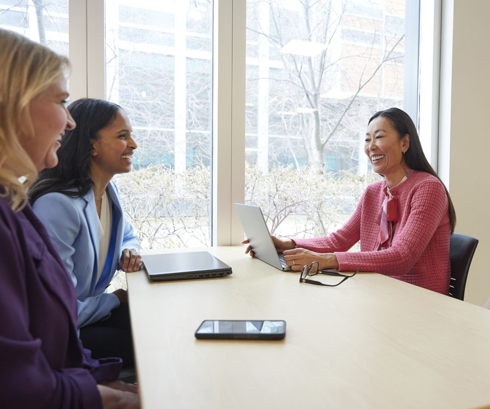 A group of three diverse women engaged in a discussion around a table, with one woman smiling while using a laptop and another holding a tablet.