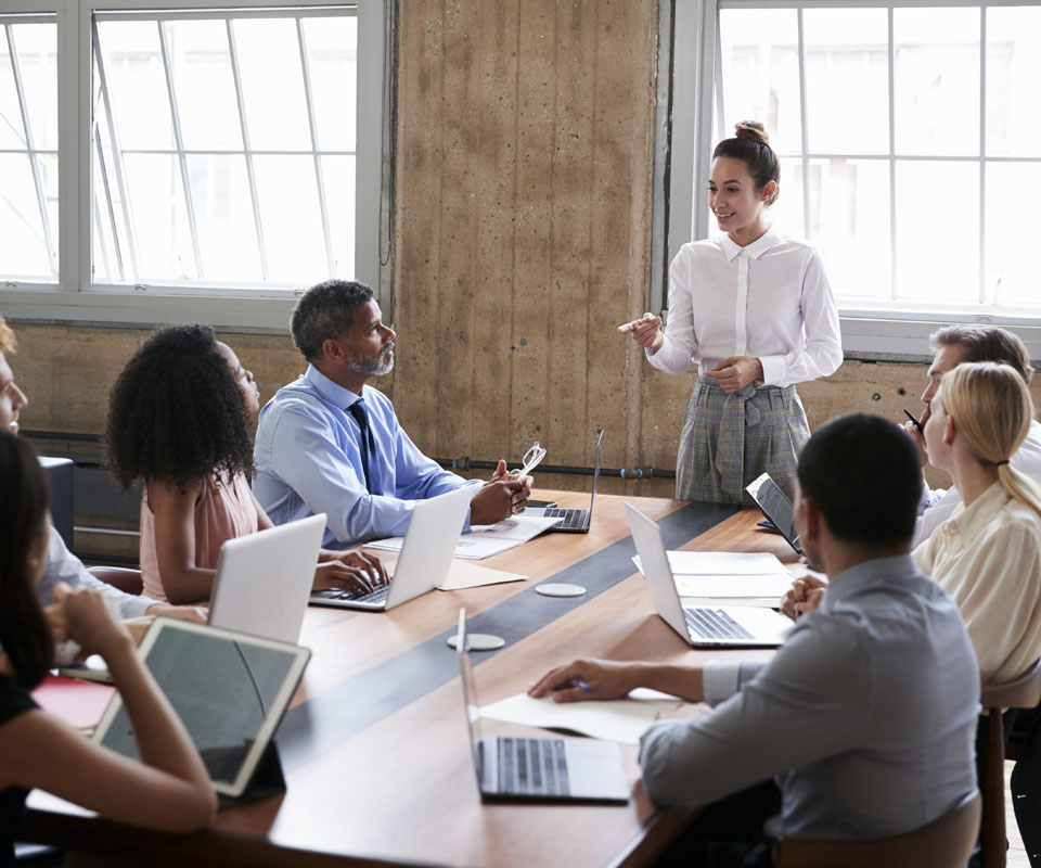 A woman presents to a diverse group of colleagues seated around a conference table, with laptops open in front of them.
