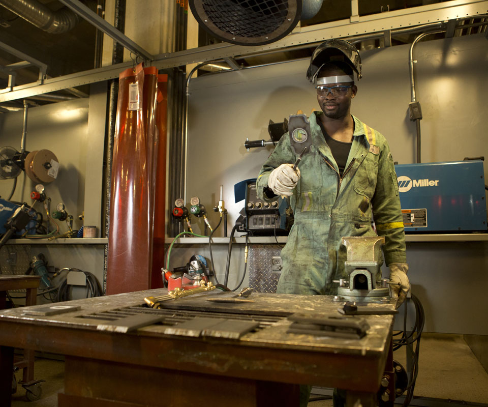 A steamfitter in protective gear stands confidently in a workshop with tools on the table and equipment in the background.