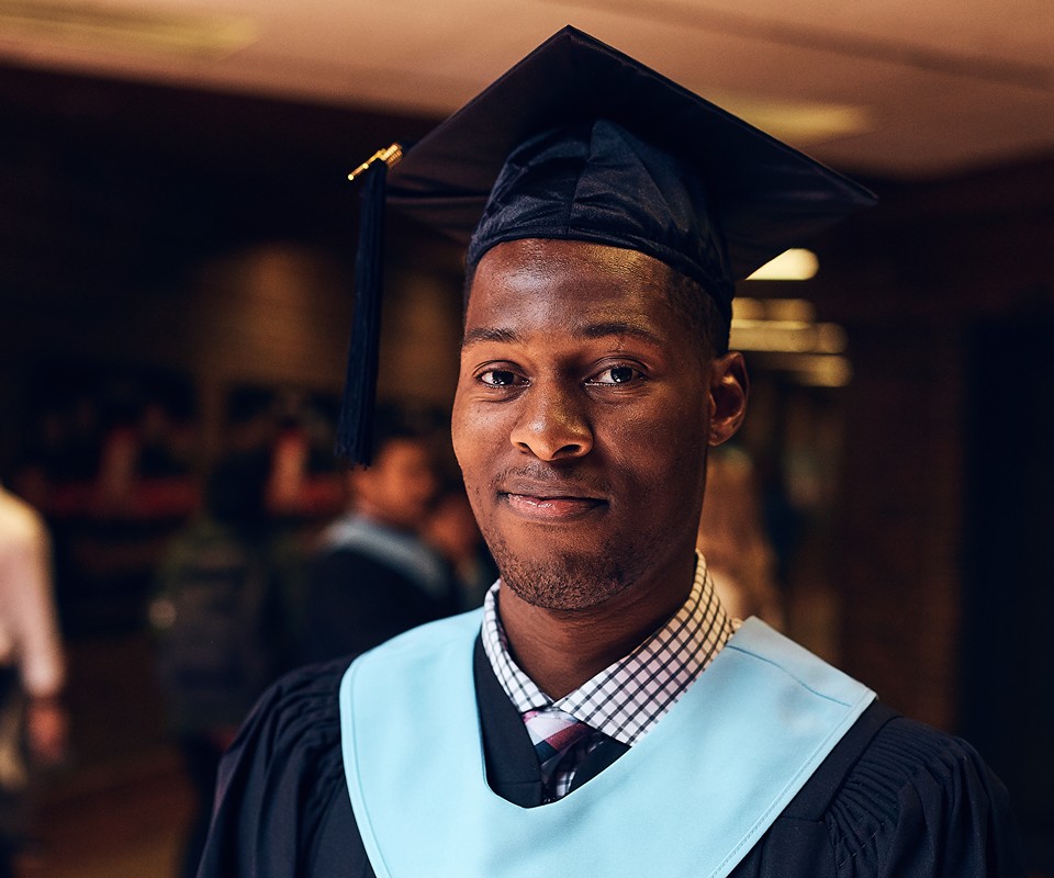 A smiling graduate in a cap and gown poses confidently, highlighting their achievement in a school hallway.