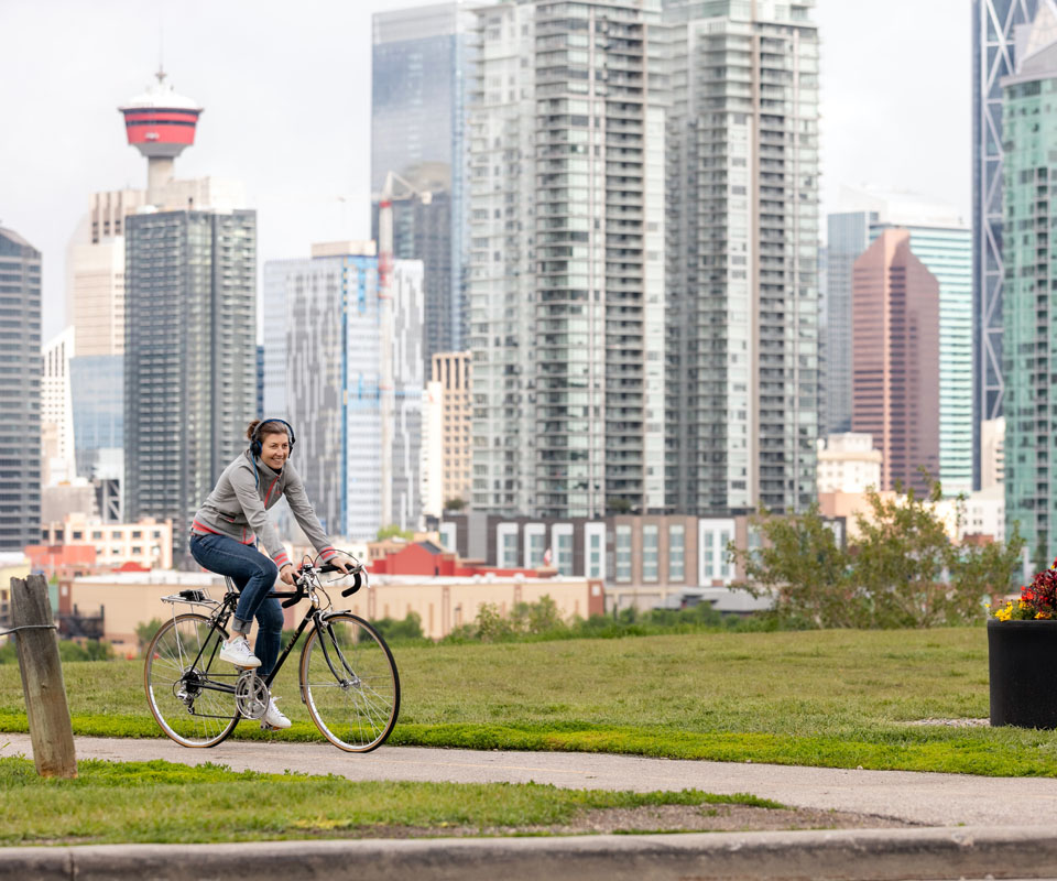A woman rides a bicycle through a green space with the downtown Calgary skyline in the background.