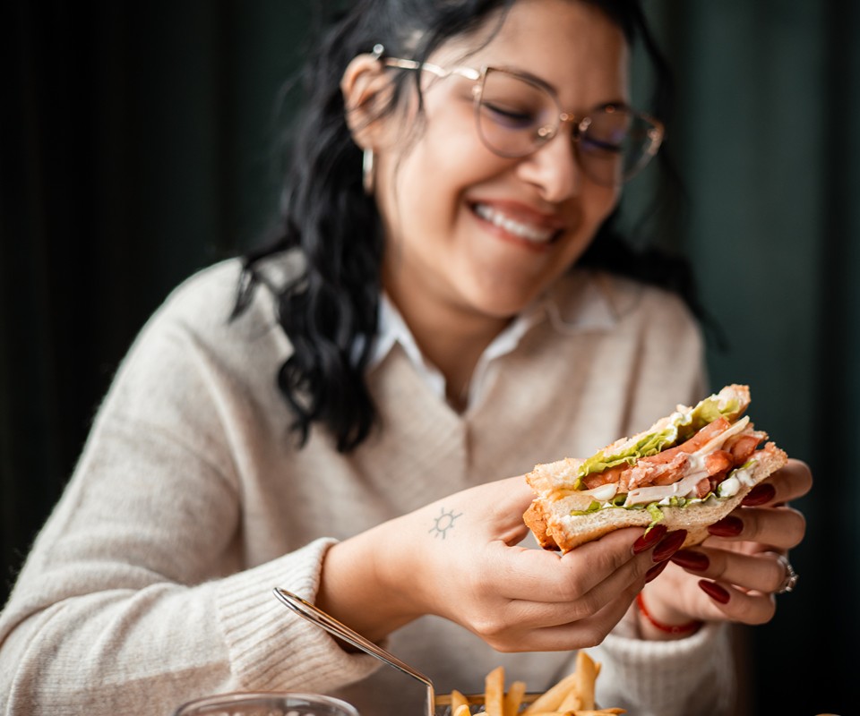 A woman smiles while holding a sandwich filled with lettuce and tomato, with a side of fries on the table.