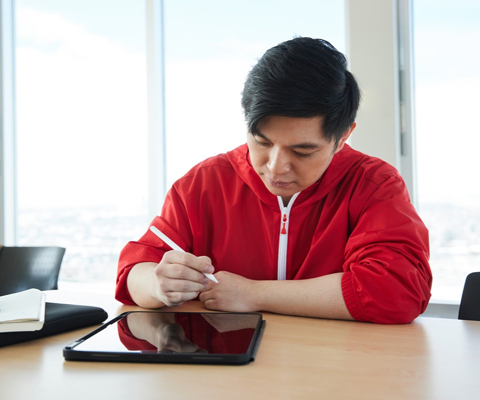 A man in a red jacket uses a stylus to write on a tablet while seated at a table in a modern workspace.